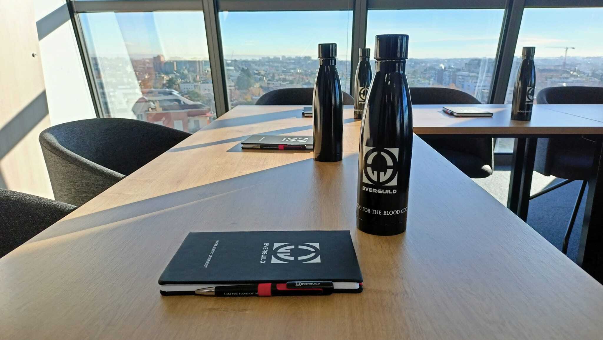 There are a few bottles and a notebook in colour black with the Everguild logo printed on them, white over black. These things are laying on a table in a bright office room.